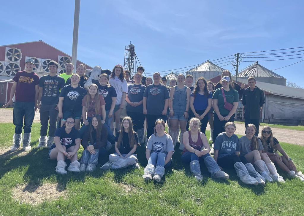 A large group of Unity Dual Credit Animal Science students posing together on a grassy area in front of a red barn and several large silver grain bins at Four Cub Farms. The students are wearing casual clothes and protective plastic boot covers for the farm tour under a clear blue sky.