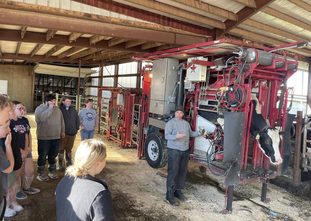 A group of Unity students, seen from behind, standing at a metal gate inside a large dairy barn. They are observing several black and white Holstein cows in a feeding area. An adult leader is standing with the group, and the barn features a high wooden-beamed ceiling.
