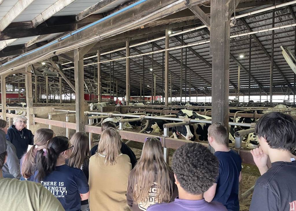 A group of Unity students, seen from behind, standing at a metal gate inside a large dairy barn. They are observing several black and white Holstein cows in a feeding area. An adult leader is standing with the group, and the barn features a high wooden-beamed ceiling.