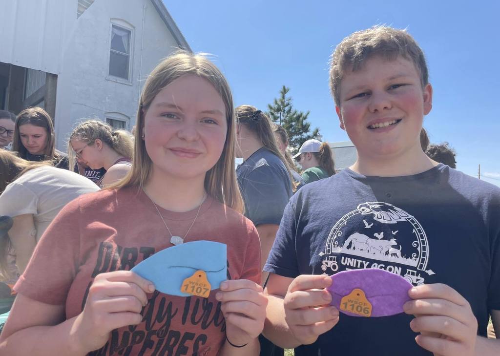 Two Unity students smiling while holding up colorful livestock ear tags during their visit to Petzel Sheep Farm. The student on the right is wearing a "Unity Ag Ed" t-shirt, and other students are visible in the background under a bright, sunny sky.