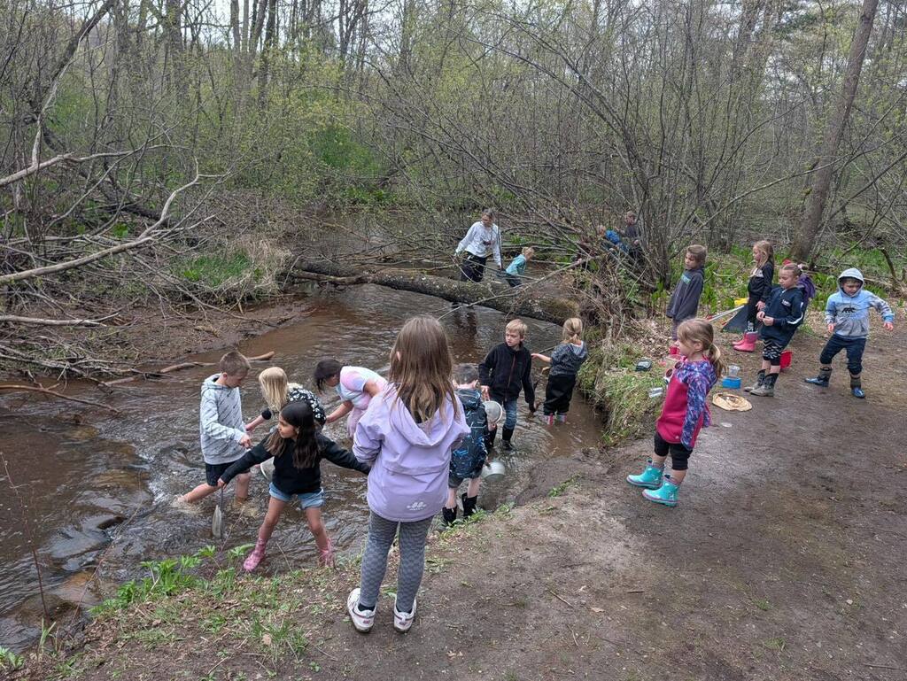 A group of first-grade students gathered outdoors around an instructor who is holding up a brown animal pelt for the children to examine.