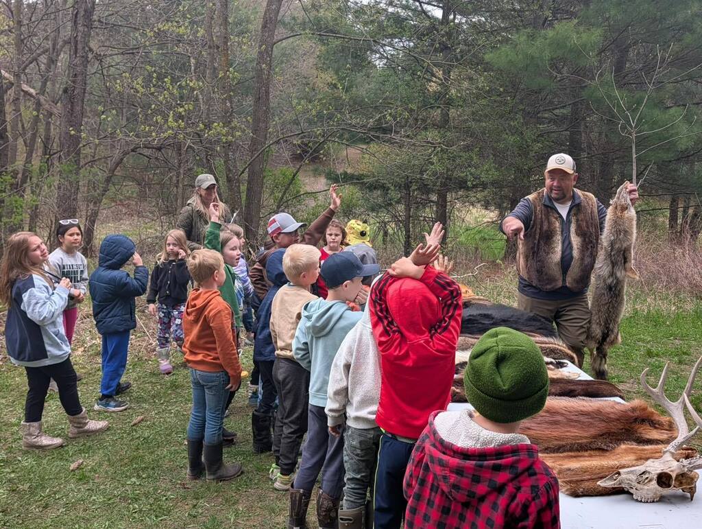 A group of young students standing in a wooded area, listening to an instructor point to several animal pelts and deer antlers displayed on a table.