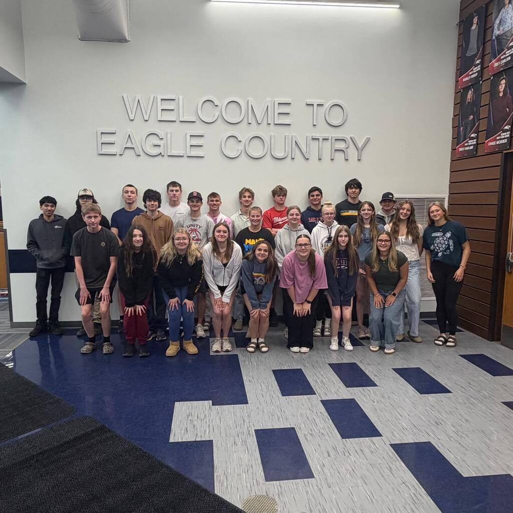 A large group of high school students posing together in the school lobby. They are standing in front of a white wall that says 'WELCOME TO EAGLE COUNTRY' in large silver letters.