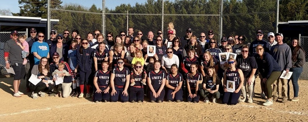 A large group of Unity softball players in their black and red uniforms posing on the dirt field with a large group of staff members and supporters standing behind them in front of a chain-link fence.