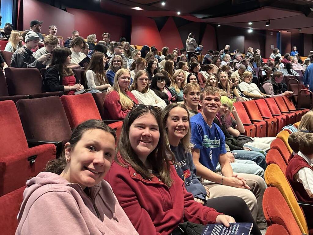 A group of Unity High School students smiling for a selfie while sitting in the red tiered seating of the Guthrie Theater. The rest of the audience and the stage set are visible in the background.