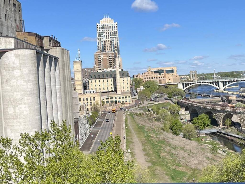A high-angle view of the Minneapolis riverfront under a clear blue sky, featuring the historic Mill City Museum grain silos, the Stone Arch Bridge in the distance, and city streets lined with green trees.