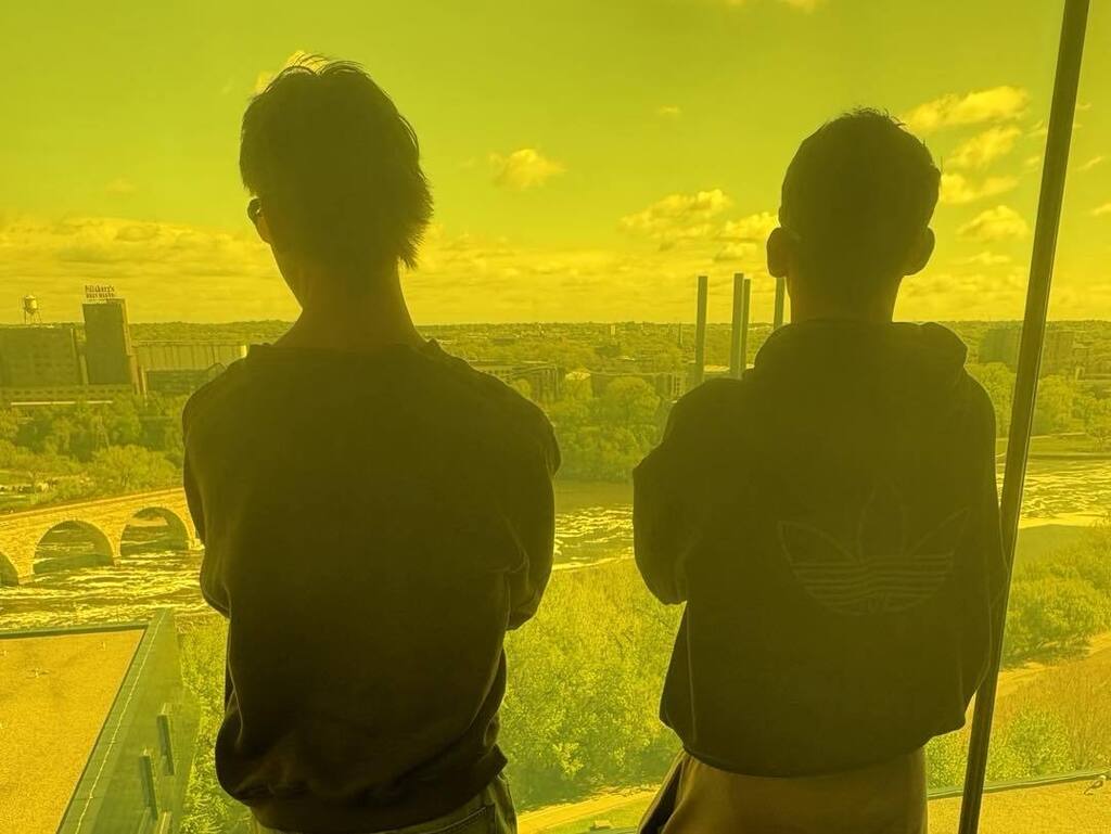 Two students seen from behind in silhouette, looking out through a bright yellow-tinted window at the Guthrie Theater. The Minneapolis skyline and river are visible through the golden hue.