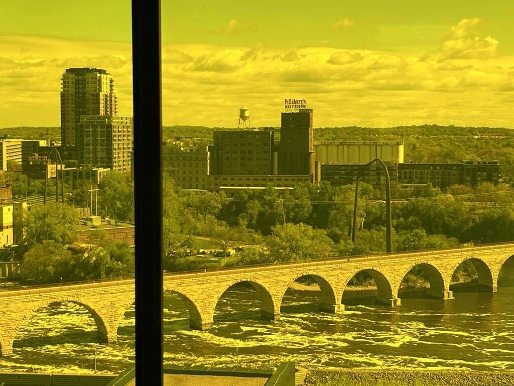A scenic view of the Stone Arch Bridge and the Minneapolis riverfront seen through a yellow-tinted glass window, creating a warm, golden monochromatic effect over the city buildings and water.