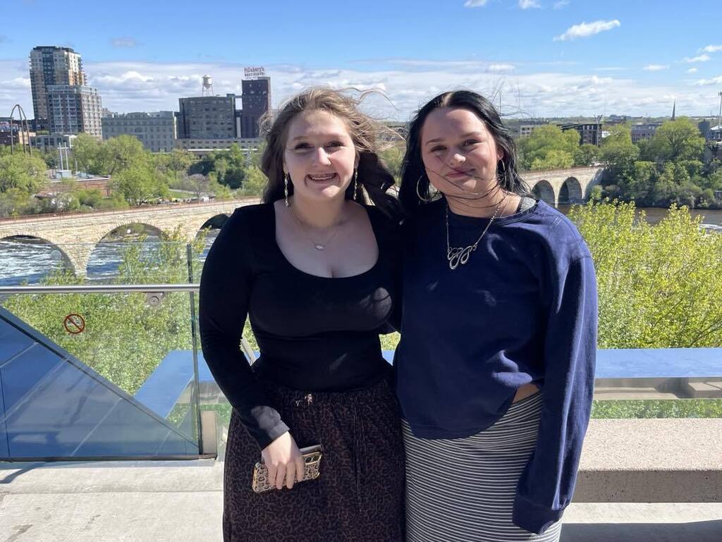 Two female AP Literature students smiling and posing together on the Endless Bridge at the Guthrie Theater, with the Stone Arch Bridge and Minneapolis skyline in the background.