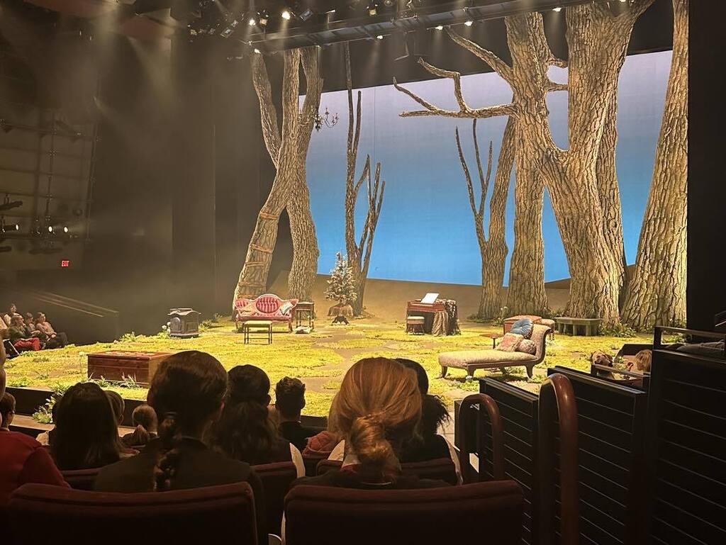 A view from the audience of the Guthrie Theater stage set for 'Little Women,' featuring large, realistic wooden tree trunks stretching to the ceiling and a cozy living room setup on a grassy stage floor.