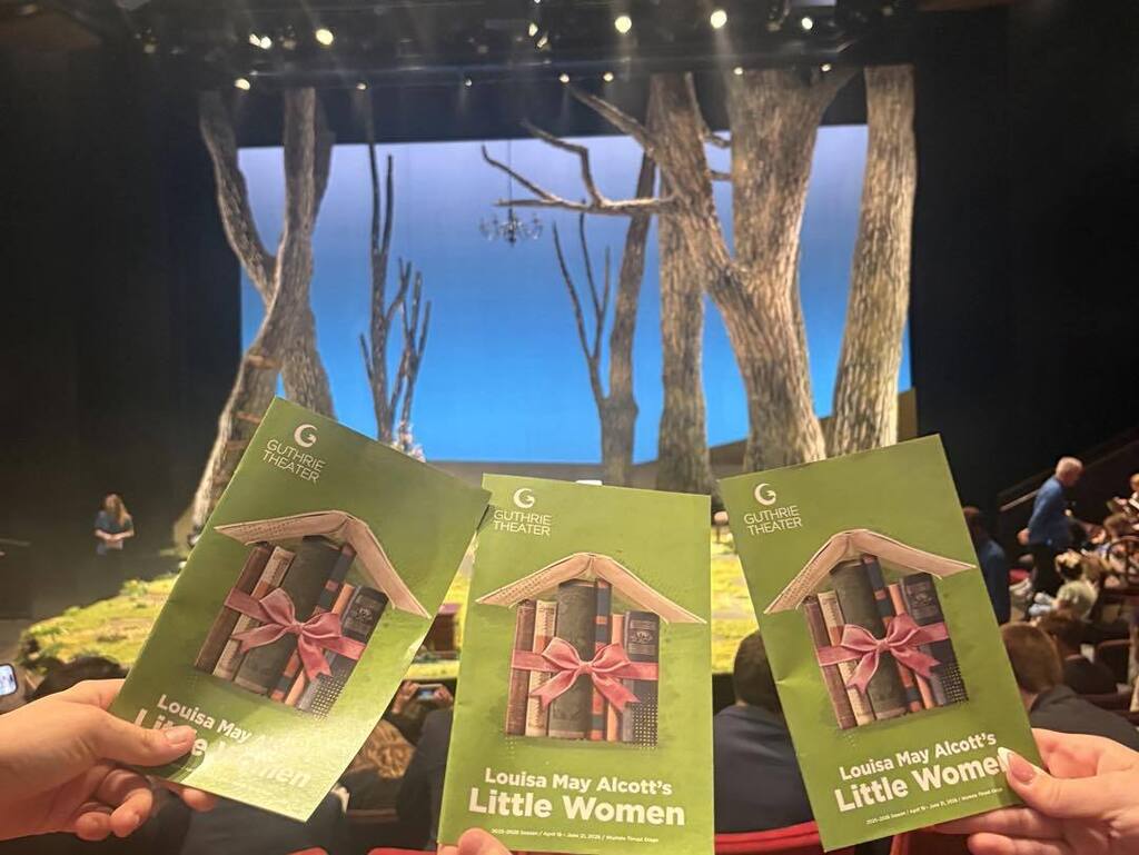 For your fourth photo featuring the playbills, here are a few alt text options for Thrillshare:  Option 1: Descriptive & Detailed (Recommended) Three green Guthrie Theater playbills for the production of 'Louisa May Alcott’s Little Women' being held up in front of the theater stage, which features large stylized trees and a blue background.