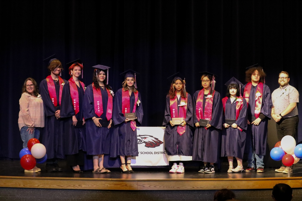 A group of Unity ADP graduates in purple caps and gowns standing on a stage with their instructors. The graduates are holding diplomas and posing in front of a blue curtain and a Unity School District banner.