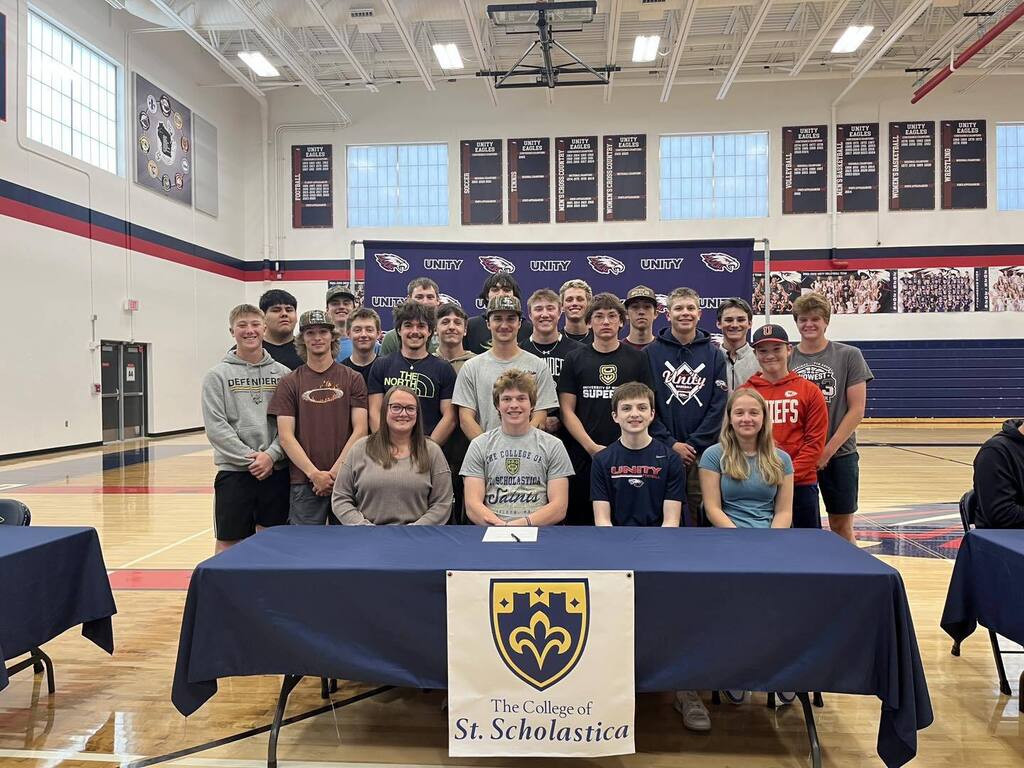 A student-athlete sits at a blue table featuring 'The College of St. Scholastica' logo and shield. He is surrounded by a large group of smiling peers and mentors in the Unity High School gymnasium.