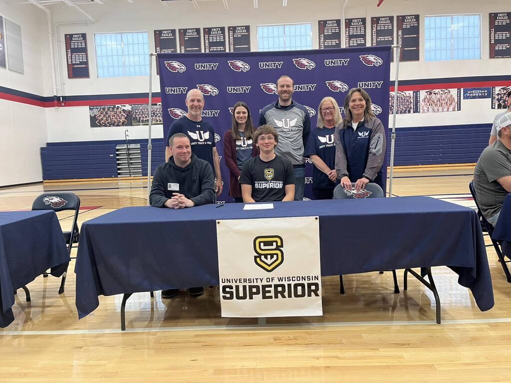 Marcus Rankin sitting at a blue signing table for the University of Wisconsin-Superior. He is joined by a group of six coaches and family members standing behind him in the Unity High School gymnasium.