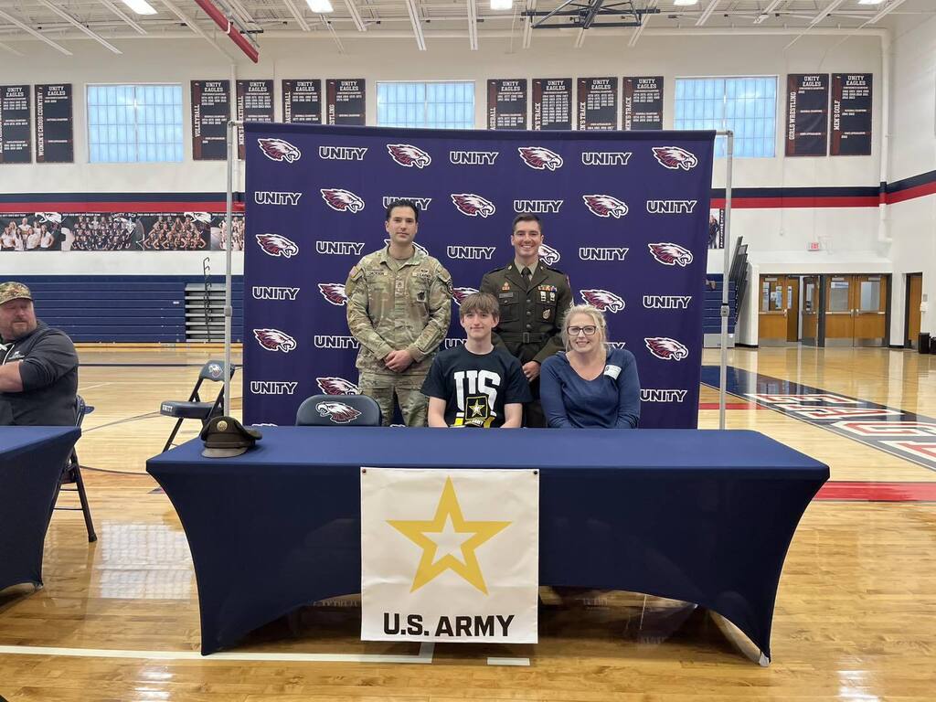 Xander Hack posing at a U.S. Army signing table, surrounded by friends and mentors.
