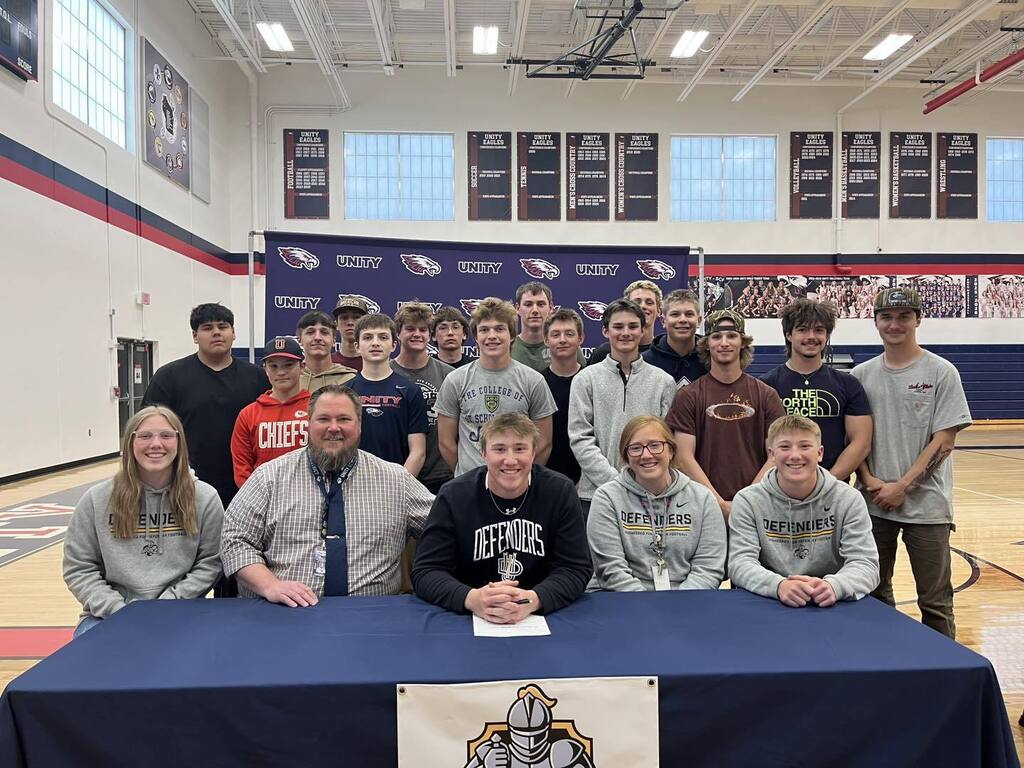 A large group of high school students and staff gathered in a gymnasium for Signing Day. A student-athlete sits at a blue table in the foreground, surrounded by peers and mentors, with Unity banners and athletic championship signs visible in the background.