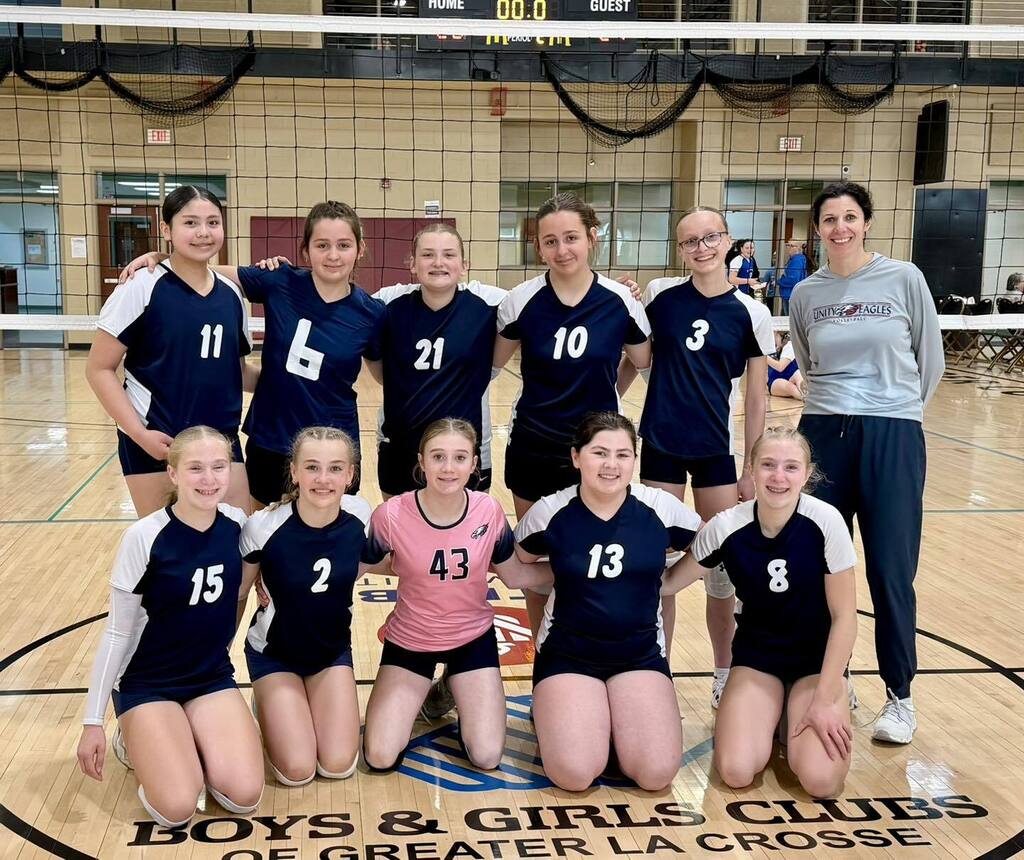 A group photo of the Unity School District 7th-grade girls' traveling volleyball team posing together on an indoor court with their coach. The nine players are wearing dark blue and pink jerseys, and their coach stands on the right. A banner at the bottom reads "Boys & Girls Clubs of Greater La Crosse."