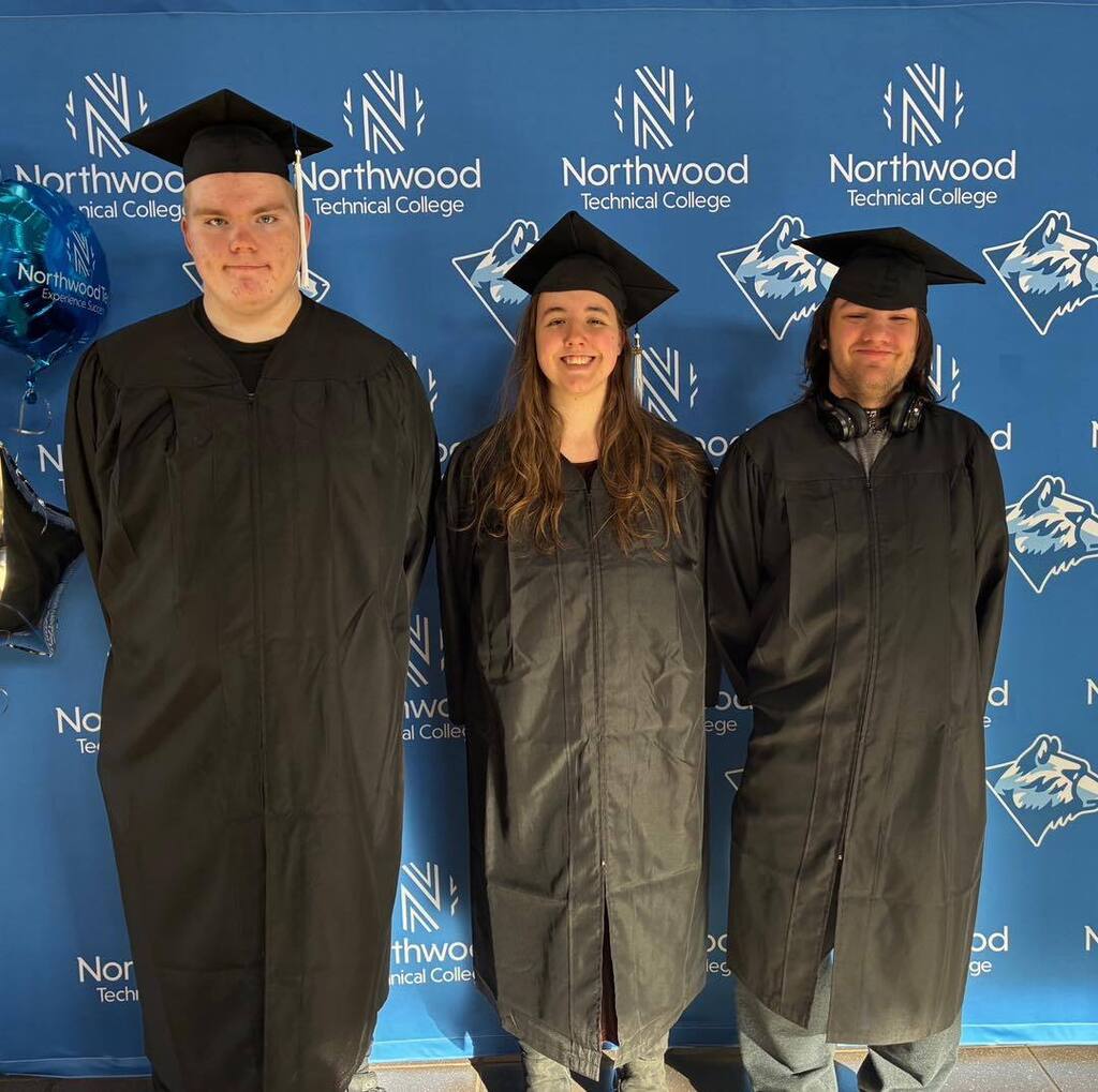 Three Unity School District graduates posing together in black graduation gowns and caps in front of a blue Northwood Technical College backdrop.
