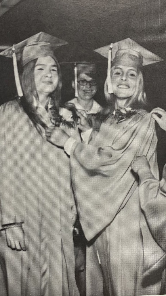 A vintage black-and-white photograph of three graduates in traditional caps and gowns adjusting their attire and smiling before a graduation ceremony.