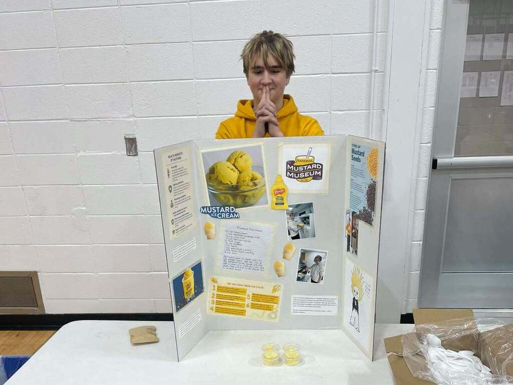 A student in a yellow hoodie stands behind a tri-fold display board about "Mustard Ice Cream" at a school capstone showcase. The board features images of ice cream, a recipe, and a National Mustard Museum logo.