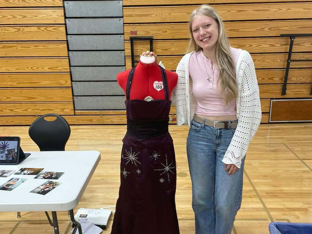 A smiling high school student stands next to her capstone project in a gymnasium: a dark purple or maroon strapless formal dress with embroidered silver starburst patterns displayed on a red dress form.
