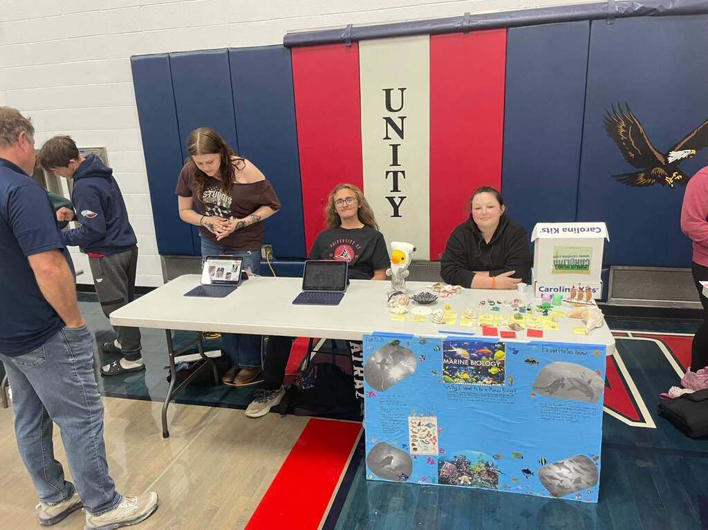 Students presenting a marine biology capstone project at a school showcase. A table wrapped in a blue poster board with marine animal photos holds displays, laptops, and student work in front of a red and black "Unity" banner in a gymnasium.