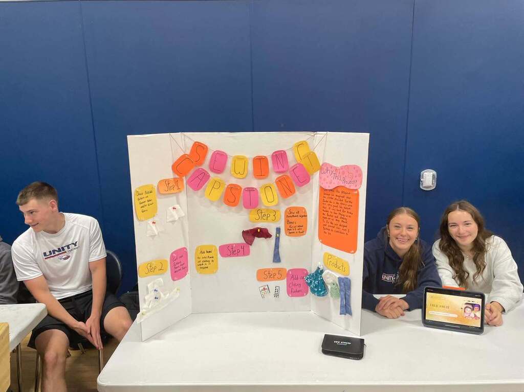 Three students smiling at a table presenting their "Sewing Capstone" project display board. The tri-fold board outlines steps 1 through 5 of their project, featuring fabric samples, colorful notes, and a tablet displaying their work.