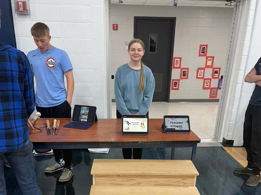 Two students presenting their capstone project at a school showcase. A table features hand-carved wooden handles, a laptop, and two tablets displaying slideshow presentations titled "Building Muscle" and "Tracking Fitness."