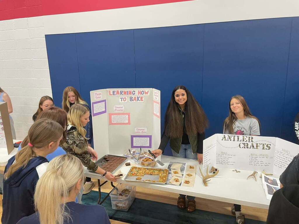 Students gathering around a project table at a school showcase. The table features two displays: a "Learning How to Bake" tri-fold board with trays of baked goods being shared, and an "Antler Crafts" display board featuring hand-crafted deer antler items.