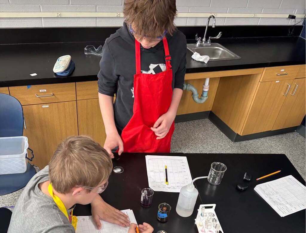 Two middle school science students wearing safety goggles and a lab apron work together at a lab table to analyze substances during a chemistry experiment.