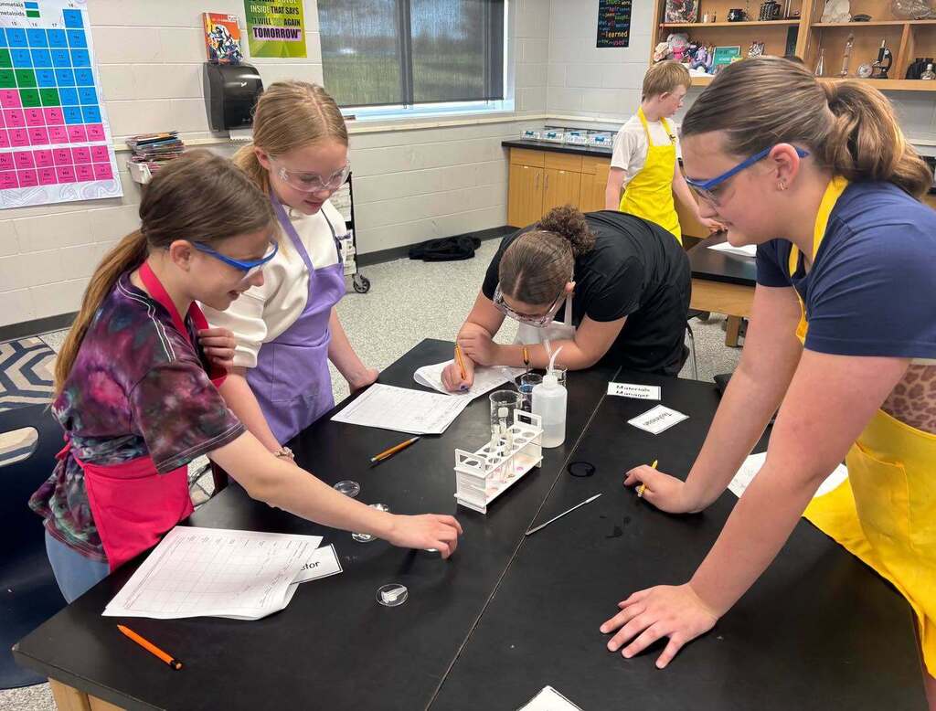 Four middle school students wearing safety goggles and lab aprons gather around a science table to collaborate on a chemistry experiment.
