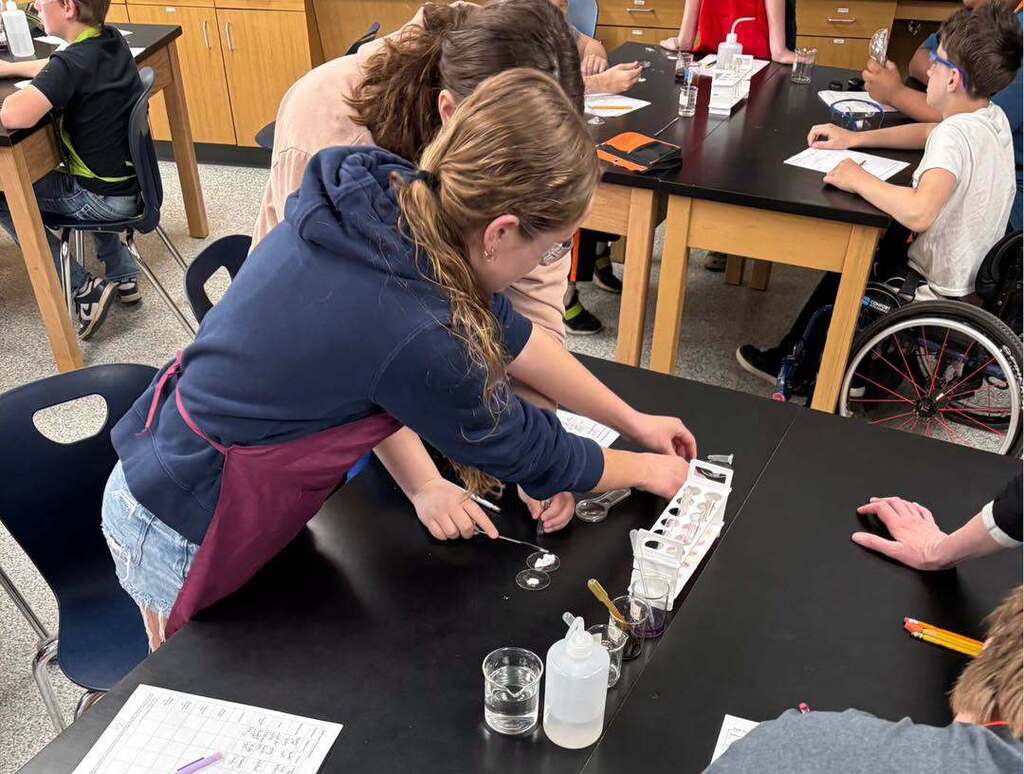 A group of middle school students, including a student in a wheelchair, wear safety goggles and work together at a lab table during a science experiment.