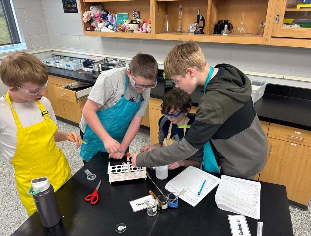 Four 6th-grade students wearing safety goggles and lab aprons work together at a science lab table to test substances using test tubes and pipettes.