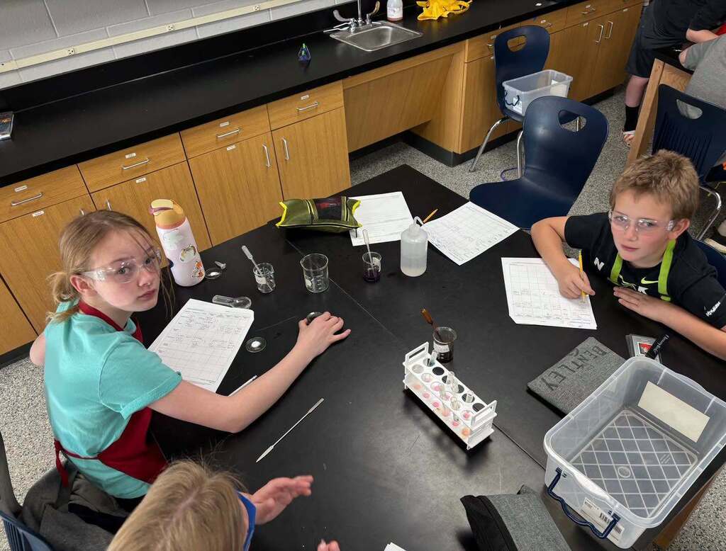 Two 6th-grade students wearing safety goggles sit at a science lab table with test tubes, worksheets, and lab materials during a forensic chemistry activity.