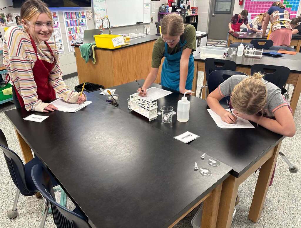 Three middle school science students wearing safety goggles and aprons work at a lab table, writing on worksheets during a chemistry experiment.