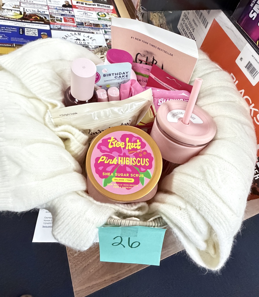 A gift basket containing Tree Hut Pink Hibiscus Shea Sugar Scrub, a pink tumbler, body care items, and books, marked with a number 26 tag.