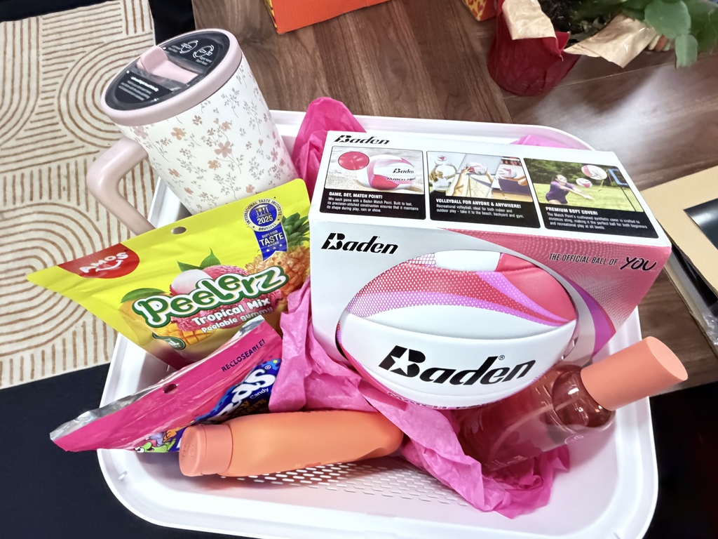 A gift basket containing a pink Baden volleyball, a stainless steel insulated tumbler, a box of Baden sports gear, a bag of Peelerz Tropical Mix candy, and other items.