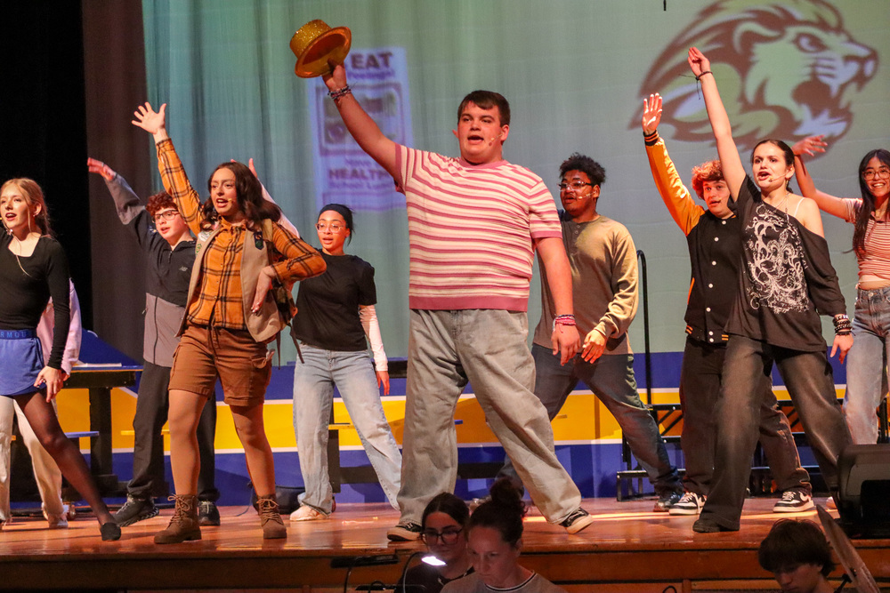 Performers on stage raising their arms, one holding a golden hat, with colorful costumes and a lion backdrop.