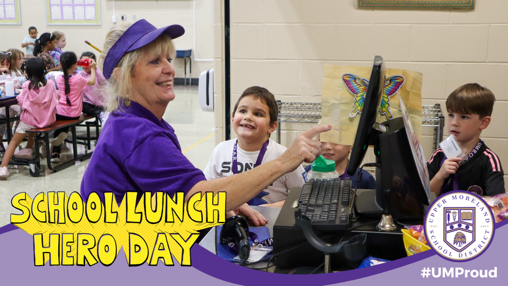 A school cafeteria scene with a woman in purple interacting with children during School Lunch Hero Day.