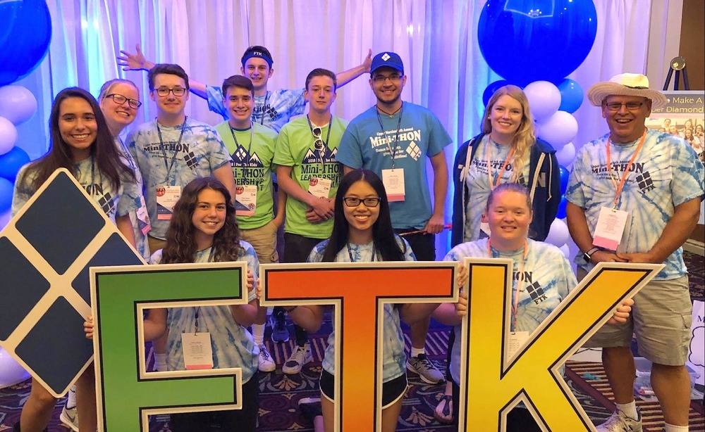 A group of people in matching tie-dye shirts pose with large letters "FTK" in front of a balloon-decorated backdrop.  