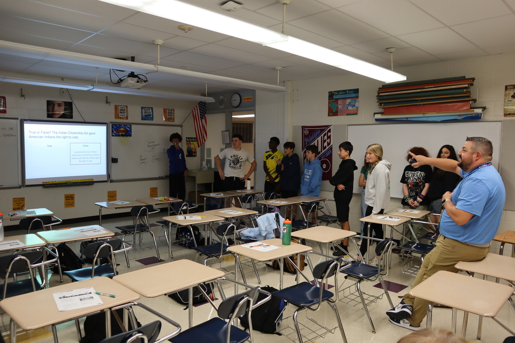 8th grade students playing an interactive true or false game. Students stand on the side of the room while the teacher stands in the middle and points at the projector screen.
