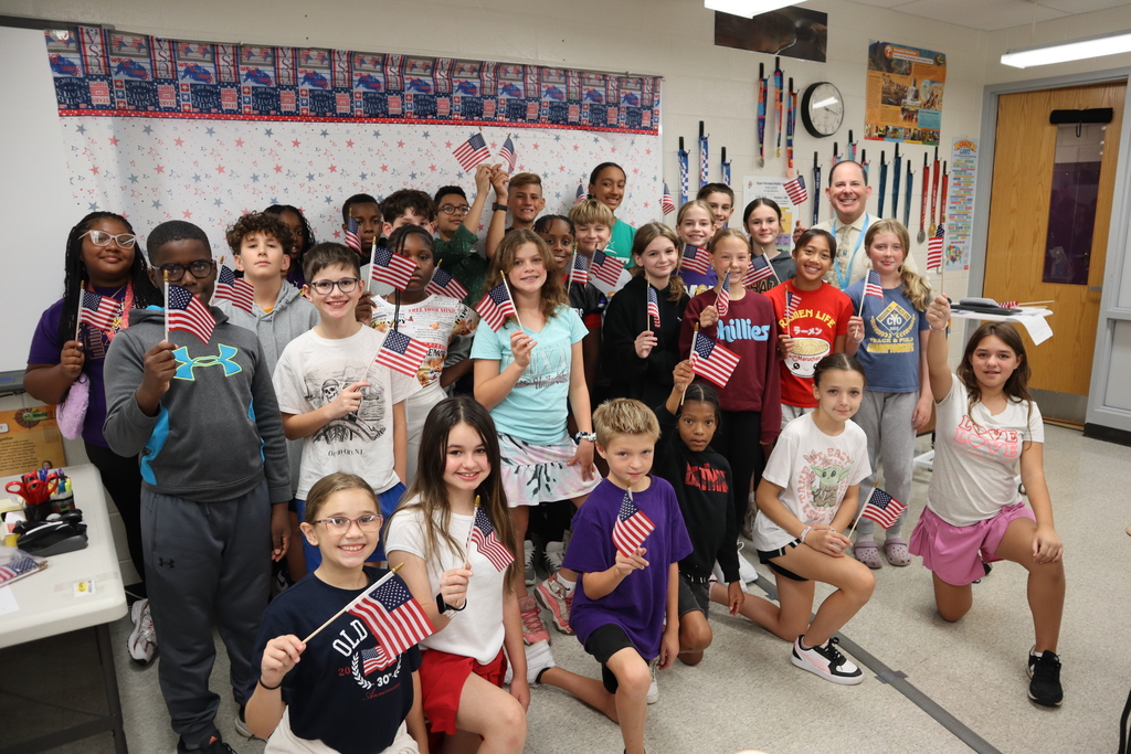 6th grade students holding an American flag and smiling as a group in front of a patriotic background.