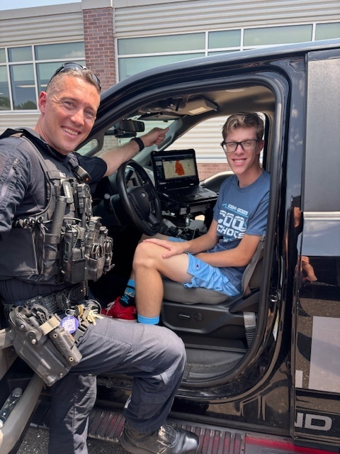 Student sits in police car and smiles with an officer