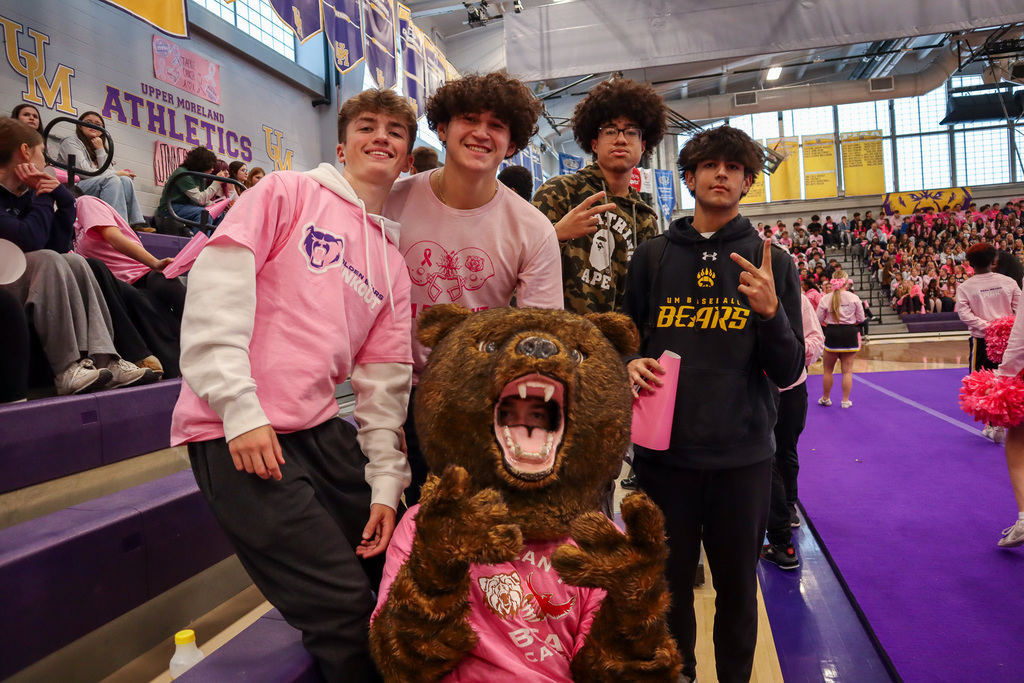 Five people posing in front of bleachers, one in a bear mascot costume, inside a school gym filled with spectators.