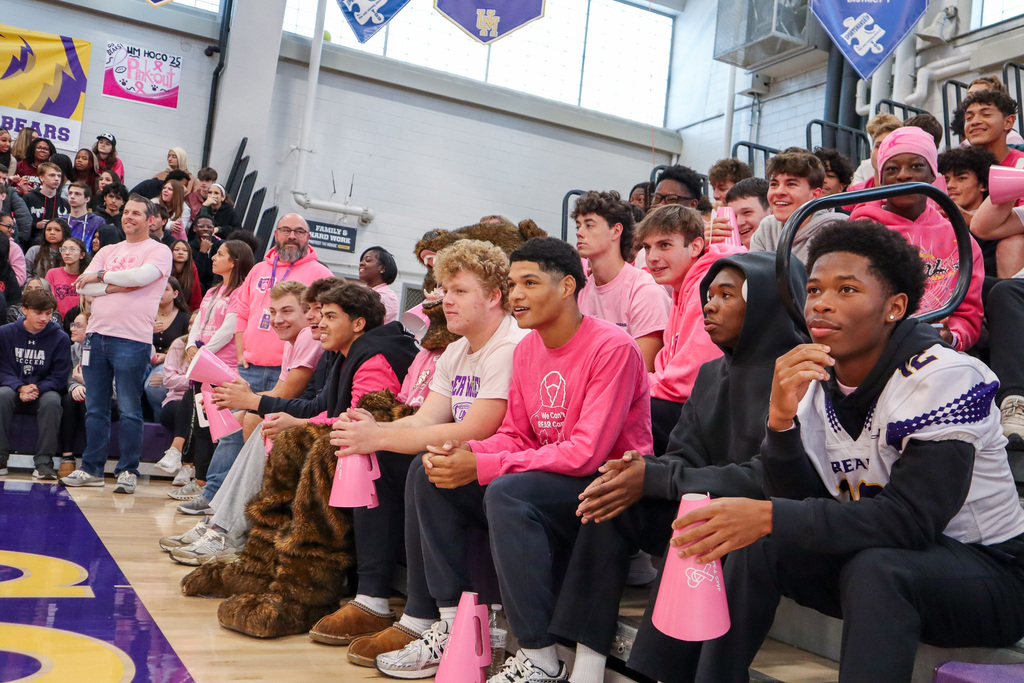 Group of students in pink clothing seated in a gym with banners and posters overhead.