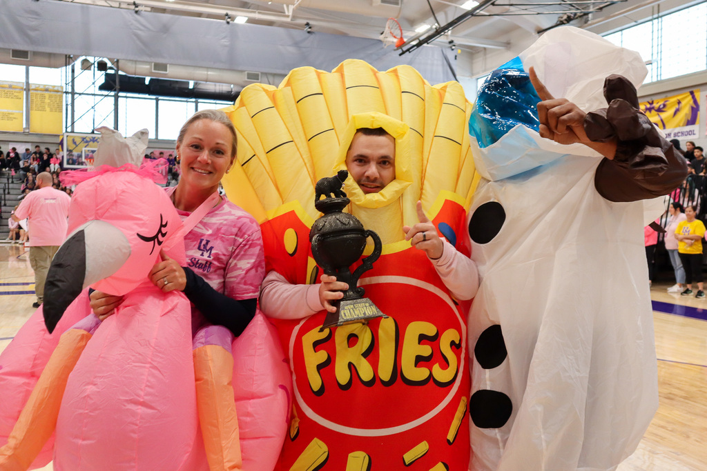 Three individuals in flamingo, fries, and snowman costumes stand in a gymnasium, with one holding a trophy.