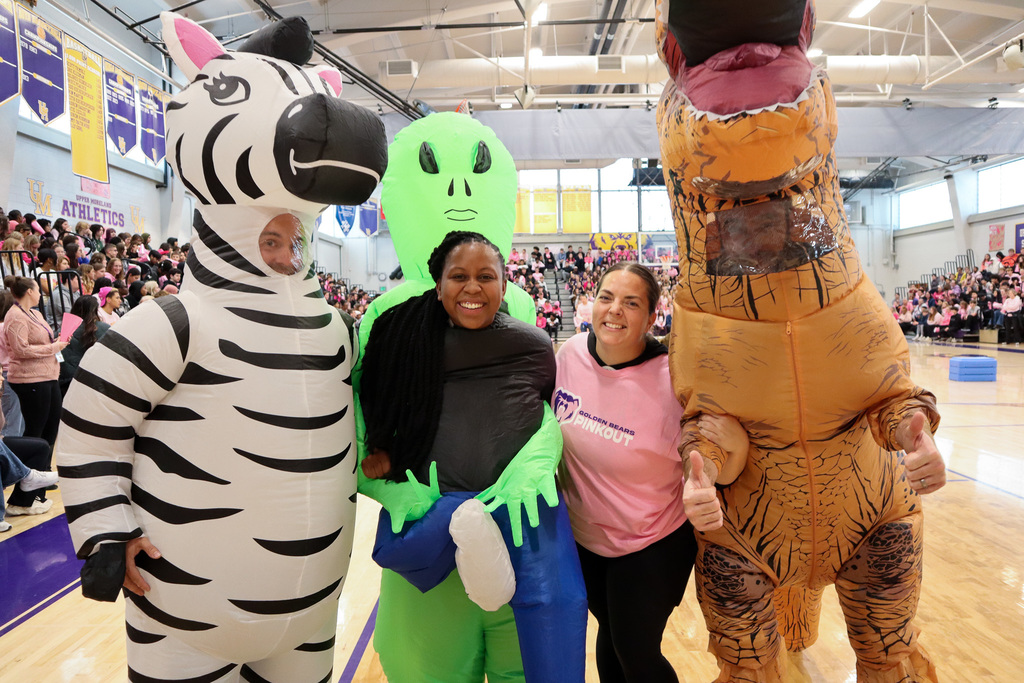 Four people in zebra, alien, and T-Rex costumes, with one wearing a pink shirt, pose in a gymnasium.