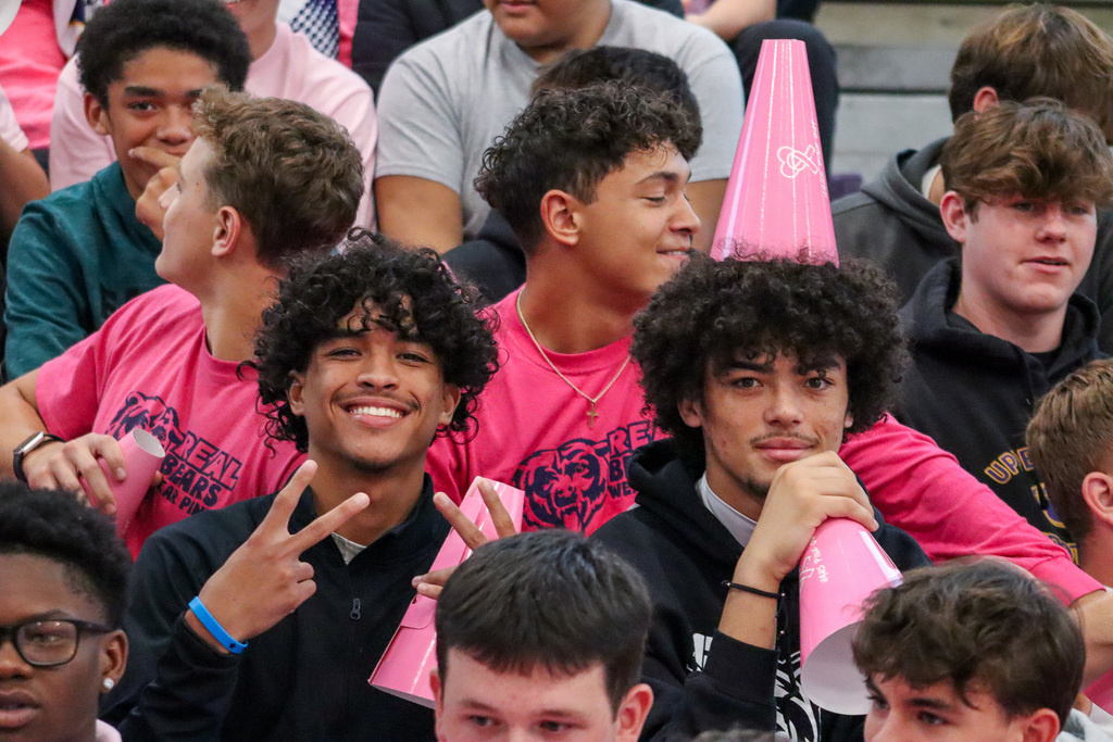 A group of young people sitting together, with two in the foreground. One smiles with a peace sign, while the other rests a pink cone on their head.