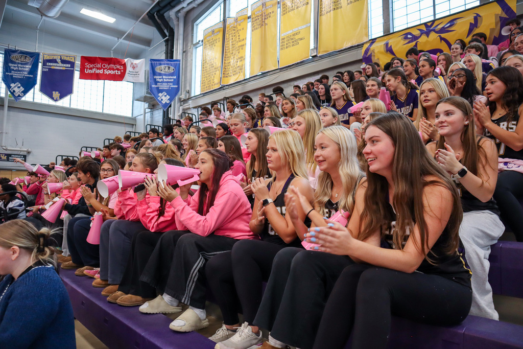 A group of spectators in a gymnasium, many wearing pink jackets and holding pink megaphones, cheering and clapping.