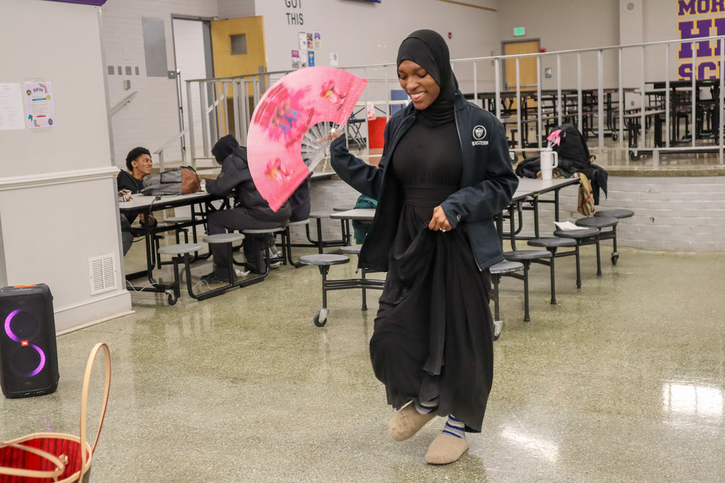Person in a cafeteria holding a pink decorative fan, wearing a black hijab and an "EASTERN" jacket. Cafeteria tables and a speaker are visible.